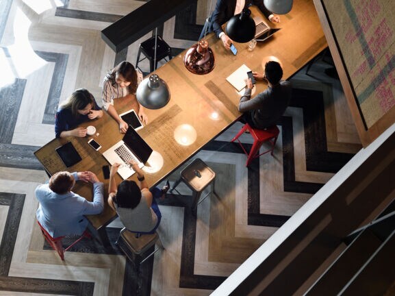 Aufsicht eines langen Holztisches, an dem mehrere junge Personen auf Metallhockern an Laptops und Smartphones arbeiten.