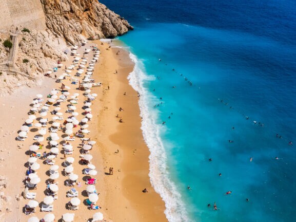 Luftaufnahme eines goldenen Sandstrandes mit Sonnenschirmen in einer Felsbucht mit Personen im türkisblauen Wasser.