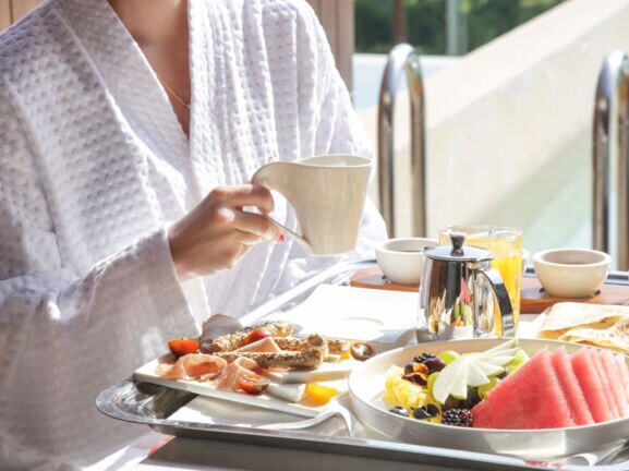 Eine Person sitzt an einem Tisch und hält eine Tasse in der Hand. Vor ihr steht ein Frühstückstablett mit Essen und Getränken.