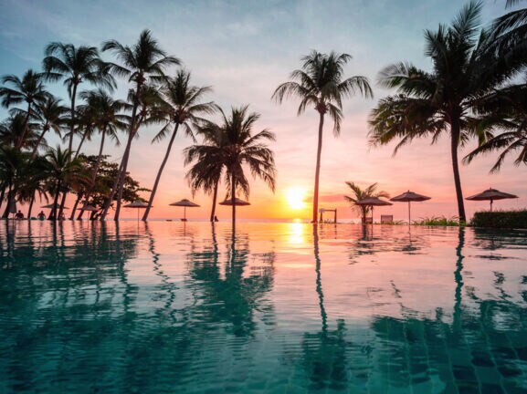 Palmengesäumter Infinity-Pool in einer Hotelanlage am Meer bei Sonnenuntergang.