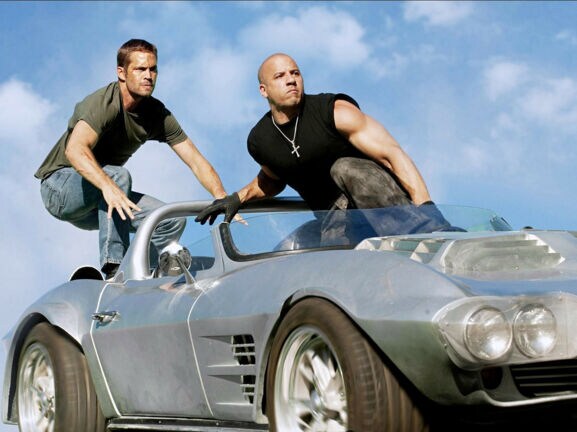 Paul Walker und Vin Diesel hocken auf einem silbernen Sportwagen ohne Verdeck.