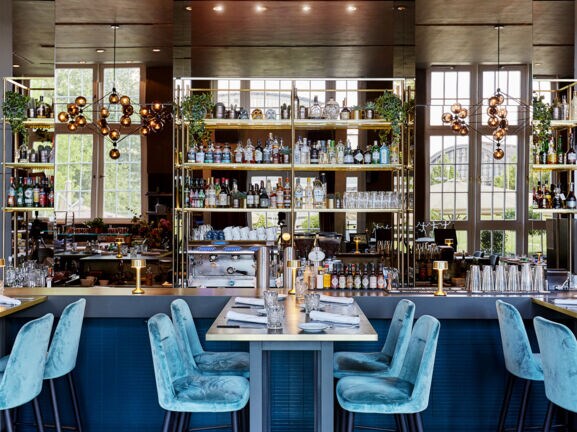 Verspiegelter Barbereich in einem modernen, edlen Restaurant mit blauen Samtstühlen an eingedeckten Tischen.