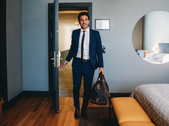 Ein Mann im Anzug trägt eine Reisetasche und betritt ein Hotelzimmer. 