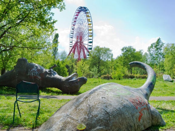Zwei umgekippte Dinosaurier-Skulpturen auf einer Wiese vor einem Riesenrad in einem Freizeitpark.
