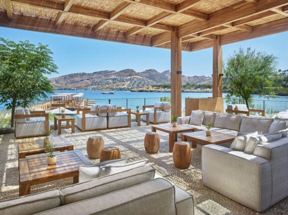 Loungebereich mit grauen Sesseln und Holztischen auf einer überdachten Terrasse an einer mediterranen Bucht.