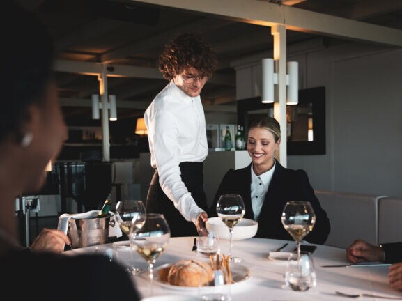 Eine lächelnde Frau wird in einem Restaurant von einem Kellner bedient, der einen Teller vor ihr auf den weiß eingedeckten Tisch stellt.
