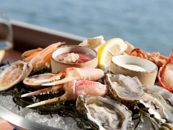 Nahaufnahme einer Platte mit gemischten Meeresfrüchten, im Hintergrund ein Glas Weißwein vor Meereskulisse.