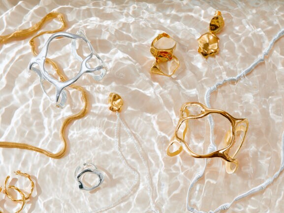 Unterschiedliche Schmuckstücke in Gold und Silber liegen auf Wasser mit Lichtspiel