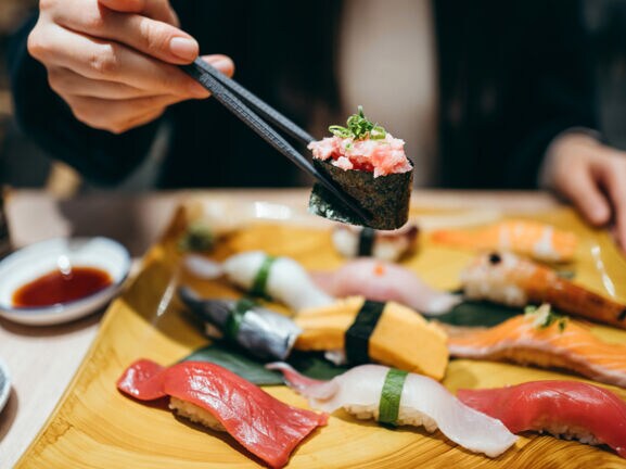 Eine Person hält eine Rolle Maki Sushi zwischen zwei Essstäbchen über einer Bambusplatte mit Nigiri auf einem Tisch in einem Restaurant.