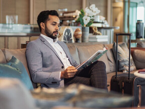 Ein elegant gekleideter Mann liest eine Zeitschrift auf einem Sofa in einer Flughafenlounge.
