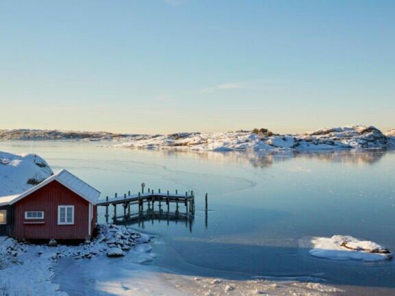 Ein rotes Holzhaus mit Steg an einem zugefrorenen See in verschneiter Landschaft in Schweden.