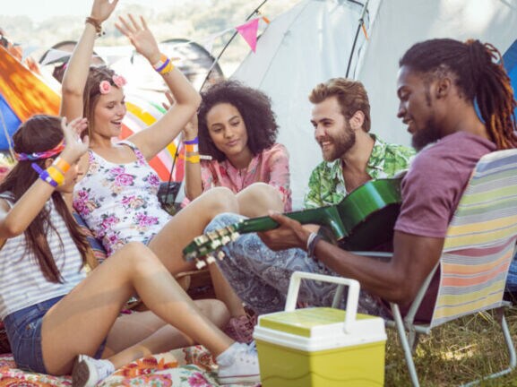 Eine Gruppe von Freund:innen sitzen zwischen Zelten, einer von ihnen spielt Gitarre.