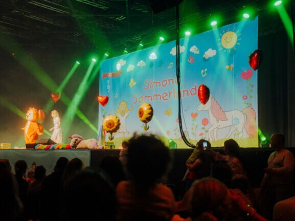 Simone Sommerland steht auf einer Bühne voller Luftballons, neben ihr eine Person im Eichhörnchen-Kostüm, vor ihr Publikum.