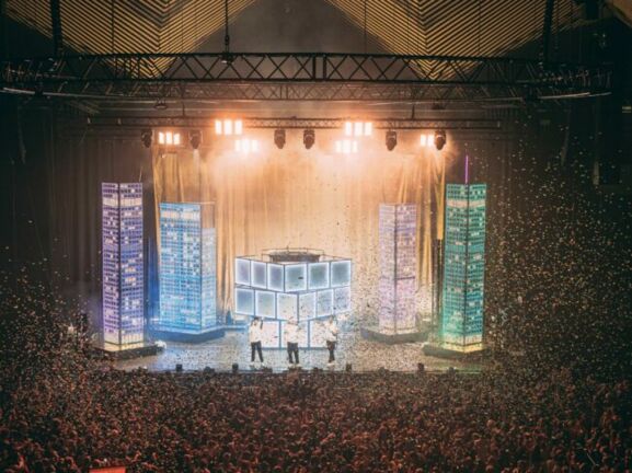 Große Menschenmenge bei einem Konzert oder Event in einer Halle mit beleuchtetem Bühnenbereich und Nebeleffekten.
