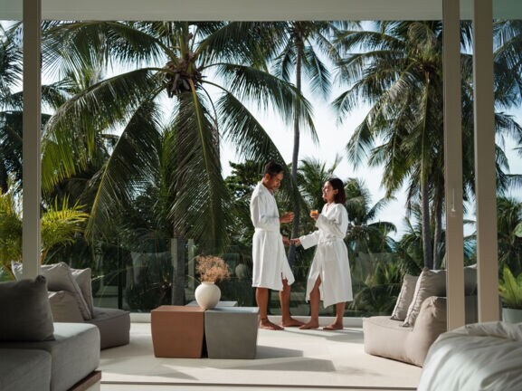 Ein glückliches Paar in weißen Bademänteln steht auf einem, von grünen Palmen umgebenen Hotelbalkon und hält ein Glas Saft in den Händen.
