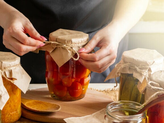 Person bindet eine Schnur um den Deckel eines Einmachglases mit eingelegtem Gemüse auf einem Holztisch.