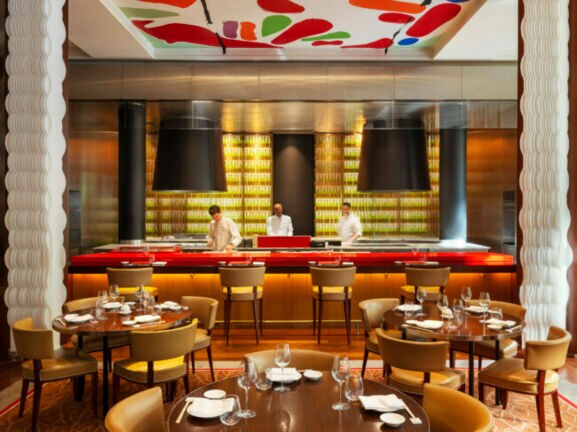 Interieur eines elegant-modernen Restaurants mir runden gedeckten Holztischen und offenem Tresen, in dem drei Köche stehen.