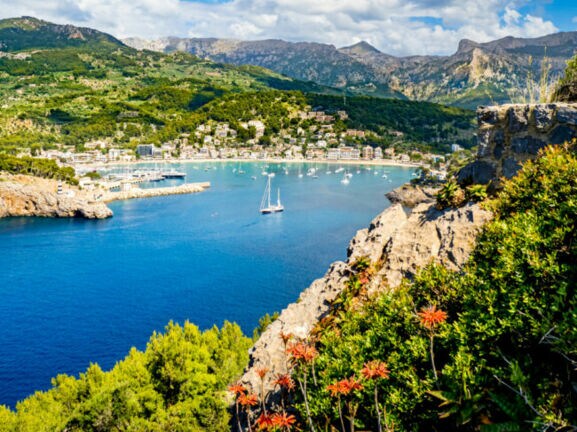 Bucht mit klarem blauem Wasser, Segelboot und Yachten, umgeben von grünen Hügeln und Bergen unter bewölktem Himmel