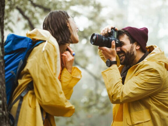 Mann mit roter Mütze und gelbem Regenmantel fotografiert lachende Frau mit blauem Rucksack und gelbem Regenmantel im Wald