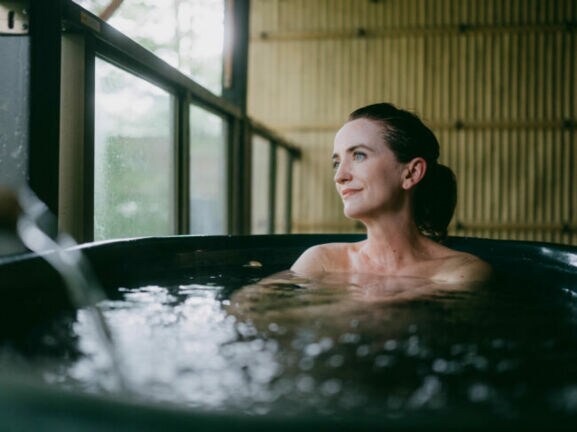 Eine Frau sitzt in einem Thermalbad aus dunklem Stein in einem japanischen Spa mit Bambuswand und schaut zufrieden durch ein Fenster hinaus.