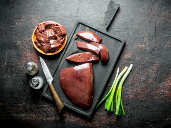 Rohe Leberstücke auf schwarzem Schneidebrett mit Messer, Frühlingszwiebeln und Gewürzstreuern auf dunklem Untergrund