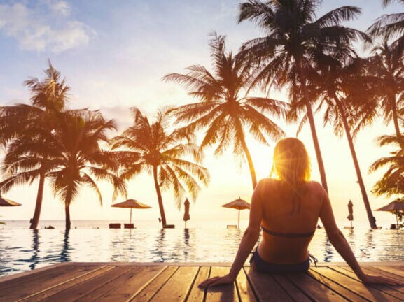 Frau sitzt am Rand eines Pools mit Blick auf Sonnenuntergang und Palmen