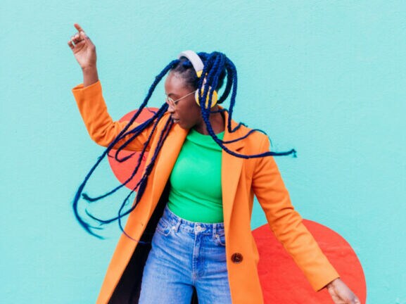 Eine junge Frau mit blauen Rastazöpfen in einem grünen Shirt und orangefarbenen Mantel tanzt mit gelben Kopfhörern vor einer türkisfarbenen Fassade.