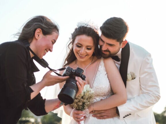 Braut in weißem Spitzenkleid mit Blumenstrauß, Bräutigam in weißem Anzug umarmt sie von hinten, Fotografin zeigt Kamera.
