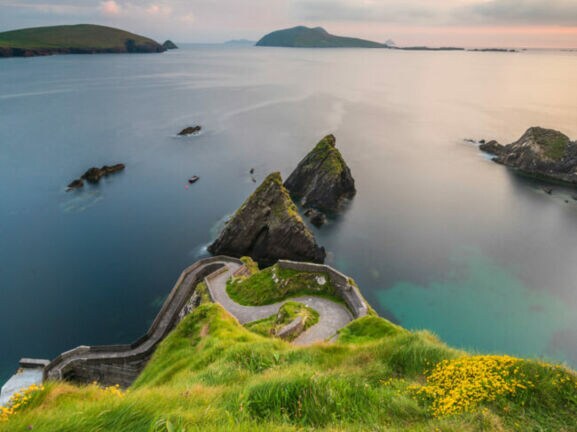 Blick auf eine Küstenlandschaft mit grünem Hügel, steilen Klippen und ruhigem Wasser. Im Hintergrund sind mehrere Inseln und ein Boot sichtbar.