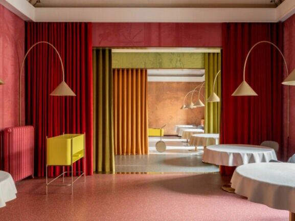 Ein eleganter Innenraum mit runden Tischen, die mit weißen Tischdecken bedeckt sind. An den Wänden hängen rote und grüne Vorhänge. Im Hintergrund sind gelbe und orangefarbene Wände sichtbar.