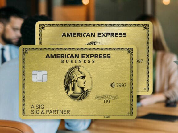 Eine Geschäftsfrau mit Klemmbrett in den Händen lehnt an einem Konferenztisch mit Personen in einem modernen Büro und schaut zufrieden in die Ferne, daneben eine Collage zweier Freisteller von goldenen Kreditkarten von American Express.