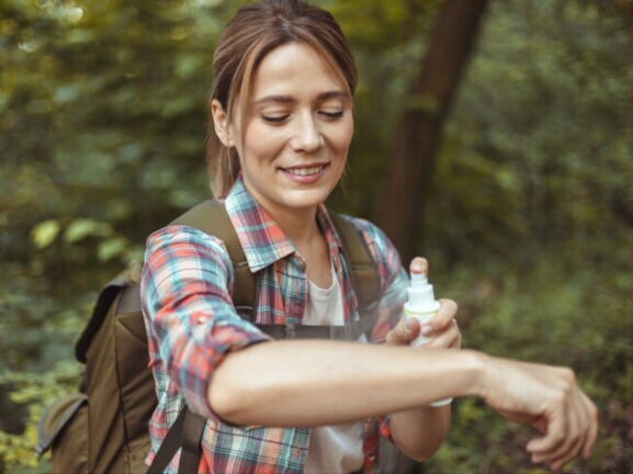 Person mit kariertem Hemd und Rucksack trägt im Wald Insektenschutzspray auf den Arm auf.