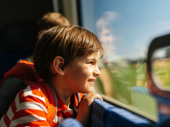 Ein Kind mit gestreiftem Oberteil lehnt sich am Zugfenster und schaut nach draußen. Im Hintergrund ist ein weiteres Kind zu sehen.