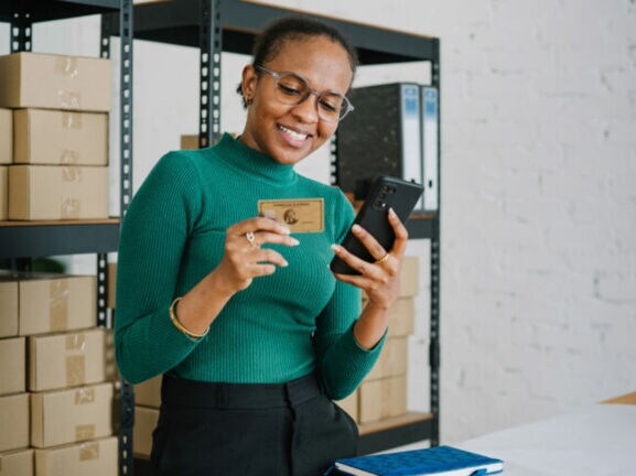 Eine lächelnde Frau im grünen Shirt steht vor einem Lagerregal mit Kartons und hält ein Smartphone sowie eine goldene Kreditkarte von American Express vor sich in den Händen.