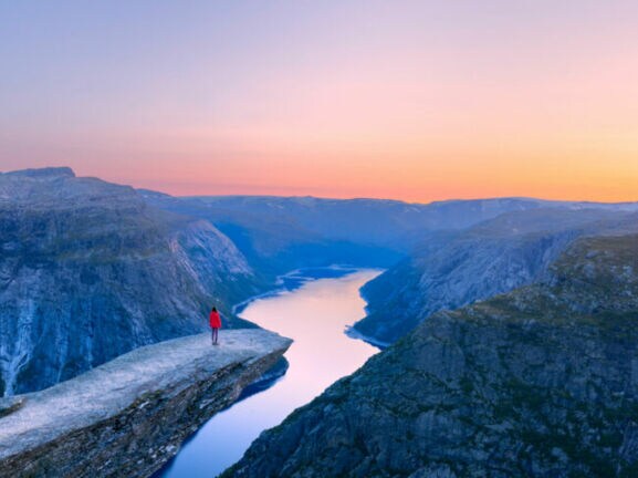 Bergklippe Trolltunga mit Person in roter Jacke, die auf dem Felsvorsprung über einem tiefen Fjord steht, umgeben von Bergen im Abendlicht.