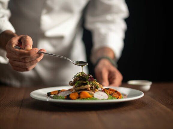 Die Hände eines Kochs, der Soße aus dem Löffel auf eine Mahlzeit auf einem weißen Teller gießt.