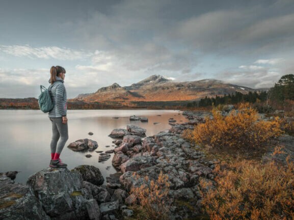 Eine Frau mit Rucksack steht auf einem Stein an einem See in flacher Felslandschaft mit herbstlicher Vegetation.