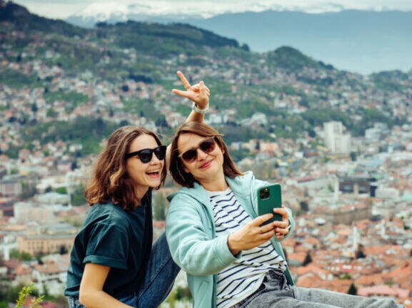 Zwei junge Frauen sitzen auf einer Mauer mit Blick auf eine Stadt, eine macht ein Selfie mit einem Smartphone und zeigt ein Peace-Zeichen.