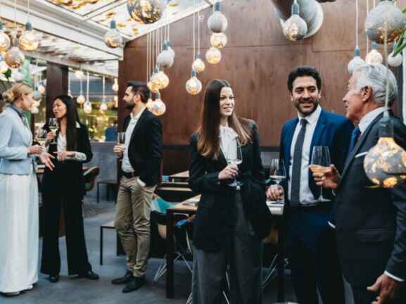 Personen in eleganter Kleidung stehen mit Weingläsern in den Händen beieinander in einem elegant geschmückten Raum.