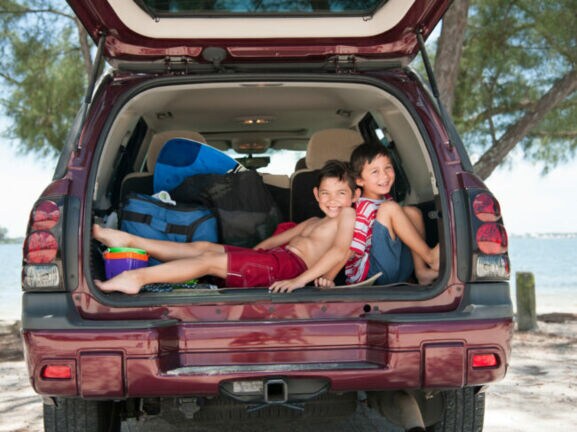 Zwei Jungen in Strandoutfits sitzen fröhlich in einem offenen Kofferraum, hinter ihnen das Meer.