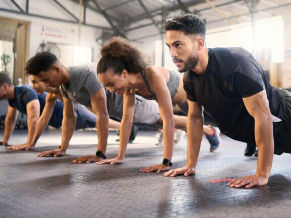 Mehrere Personen in Sportkleidung machen Liegestütze in einem hellen Fitnessstudio mit Gummiboden.