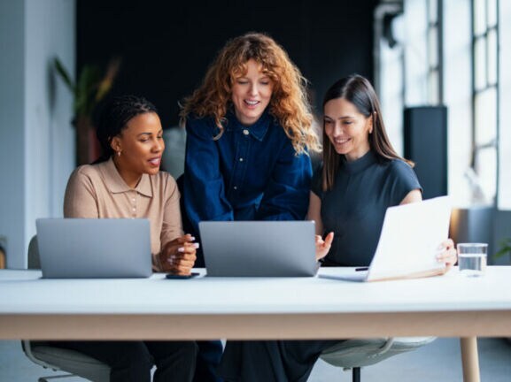 Drei Frauen schauen gemeinsam auf ein Laptop auf einem großen Tisch mit zwei weiteren Laptops in einem modernen Büro.