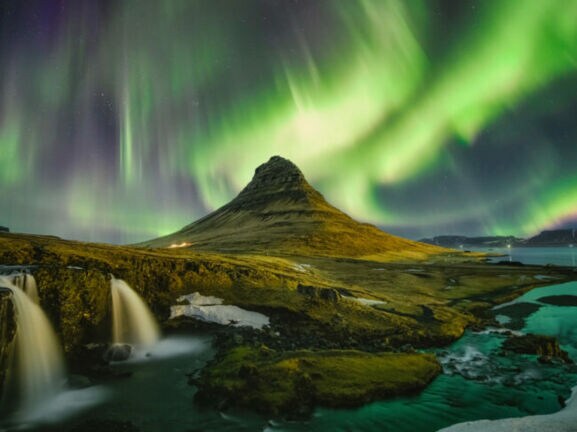 Moosbedeckte Vulkanlandschaft mit Wasserfällen unter Nachthimmel mit grün und lila leuchtenden Polarlichtern.