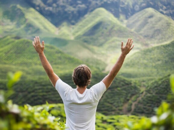 Mann mit weißen T-Shirt steht mit erhobenen Armen vor grünen, bewaldeten Hügeln.
