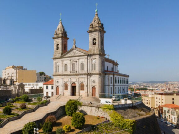 Kirche mit Zwillingstürmen auf einem Hügel über den Dächern einer südländischen Großstadt unter blauem Himmel.