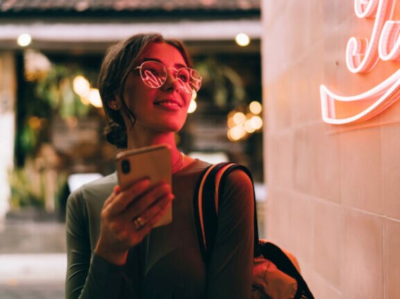 Eine junge Frau hält lächelnd ein Smartphone vor sich in der Hand, in ihrer Brille spiegelt sich der Schriftzug eines roten Logos an einer Wand.