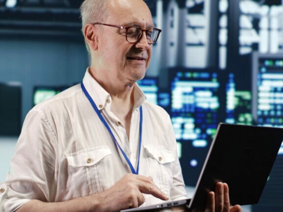 Ein Mann mit Brille ist in einem Rechenzentrum und schaut auf einen Laptop.