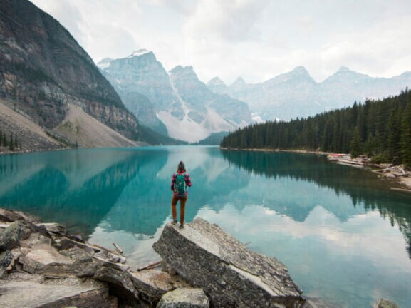 Person mit rotem Rucksack steht auf Felsen am Ufer eines klaren Bergsees, umgeben von Nadelwald und hohen Bergen.