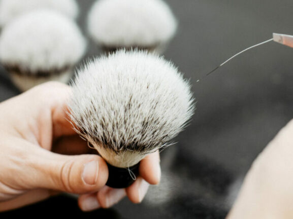 Hand hält einen weißen Rasierpinsel mit dunklen Borsten, eine zweite Hand führt eine Pinzette an die Borstenspitze.