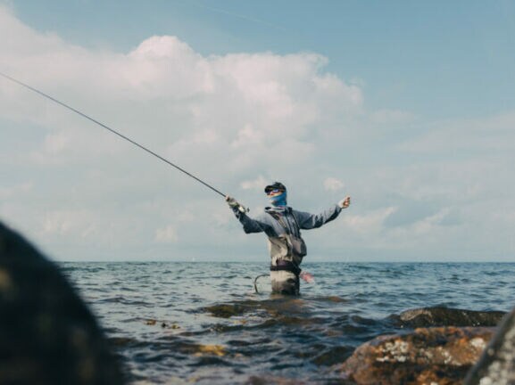 Angler steht hüfttief im Wasser zwischen Felsen und wirft eine Angelrute aus, bewölkter Himmel im Hintergrund.